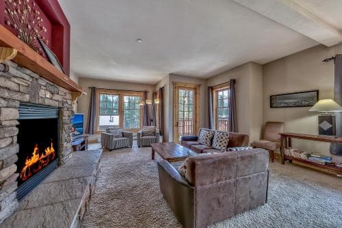 A cozy living room with a roaring stone fireplace, plush seating, and large windows inviting natural light, creates an inviting atmosphere ideal for a relaxing getaway or family vacation rental.