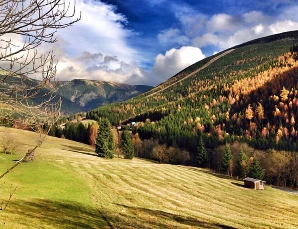 A serene mountain valley with lush green fields, autumn-colored trees, and distant peaks under a partly cloudy sky offers an ideal destination for nature lovers seeking tranquility and scenic beauty.