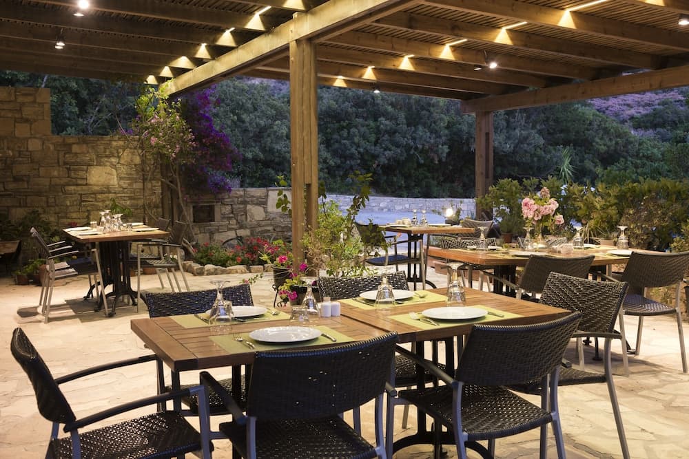 A cozy outdoor dining area under a wooden pergola with stone flooring, surrounded by lush greenery and vibrant flowers, creates an inviting atmosphere perfect for romantic dinners or intimate gatherings.