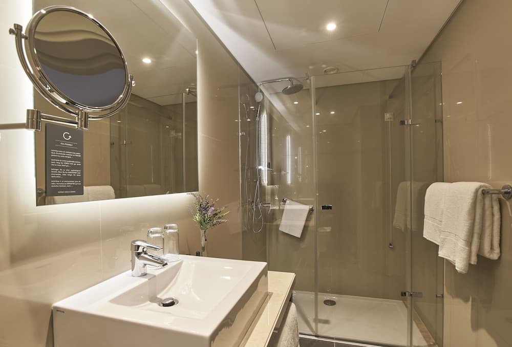 A modern, minimalist hotel bathroom features a sleek sink, a glass-enclosed shower, soft lighting, and neatly hung towels, ideal for travelers seeking comfort and cleanliness.