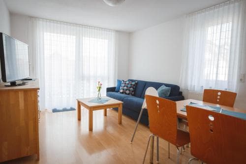 A cozy apartment living room with wooden floors, a blue sofa, dining chairs, and large windows draped in sheer curtains, ideal for travelers seeking a comfortable and modern short-term stay.