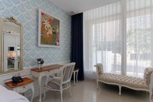 A cozy hotel room features elegant furnishings like a tufted bench, striped chair, and ornate mirror against a blue patterned wall, creating a serene and inviting atmosphere perfect for travelers seeking comfort and style.