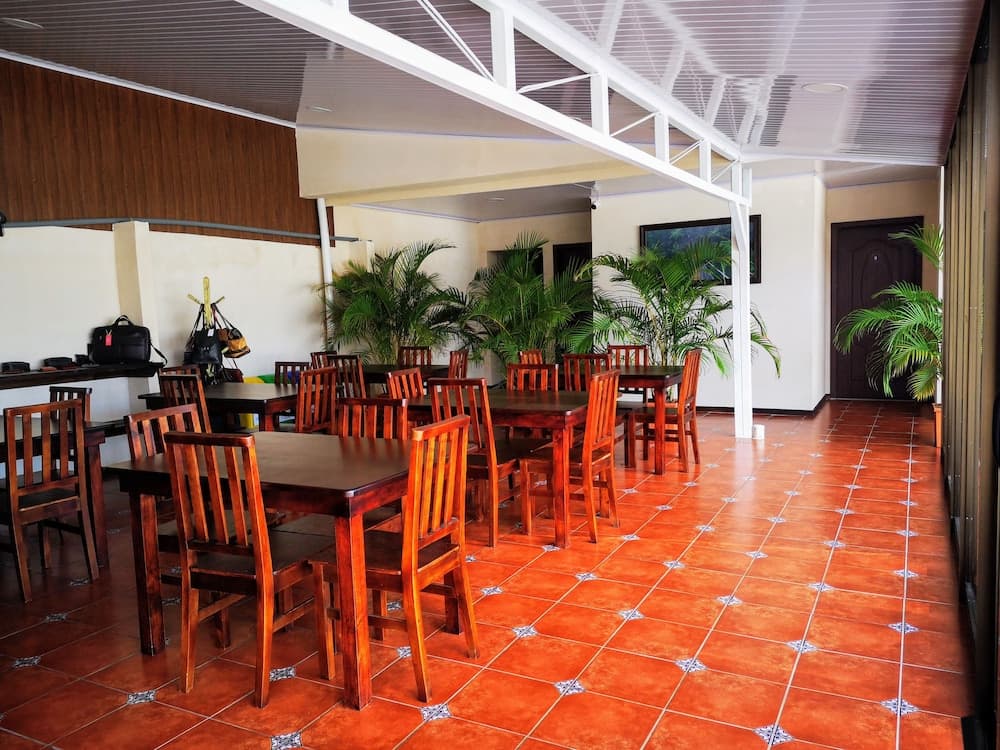 A cozy indoor dining area with wooden tables and chairs, vibrant terracotta tiles, lush potted plants, and a high-beamed ceiling, ideal for travelers seeking a relaxed meal spot in a tropical setting.