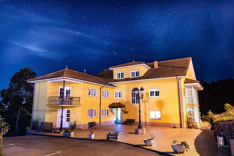 A charming yellow apartment building under a starry night sky in Asturias, Spain, offers travelers a cozy retreat with its inviting courtyard, benches, and warm lighting perfect for a serene getaway.