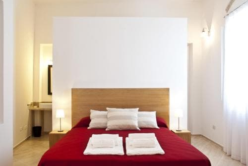 A cozy hotel room features a neatly made bed with red linens, soft lighting from bedside lamps, and a minimalist design, ideal for travelers seeking comfort and simplicity.