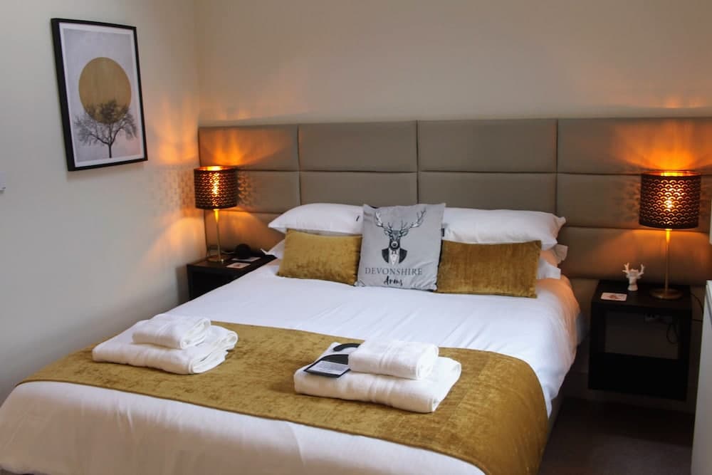 A cozy hotel room features a neatly made bed adorned with golden accents and a "Devonshire Arms" pillow, flanked by warm-lit lamps and framed artwork, ideal for travelers seeking comfort and relaxation.