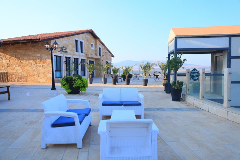 A serene outdoor patio features white wicker furniture with blue cushions, potted plants, and a stone building under a clear sky, ideal for relaxing at a tranquil resort destination.
