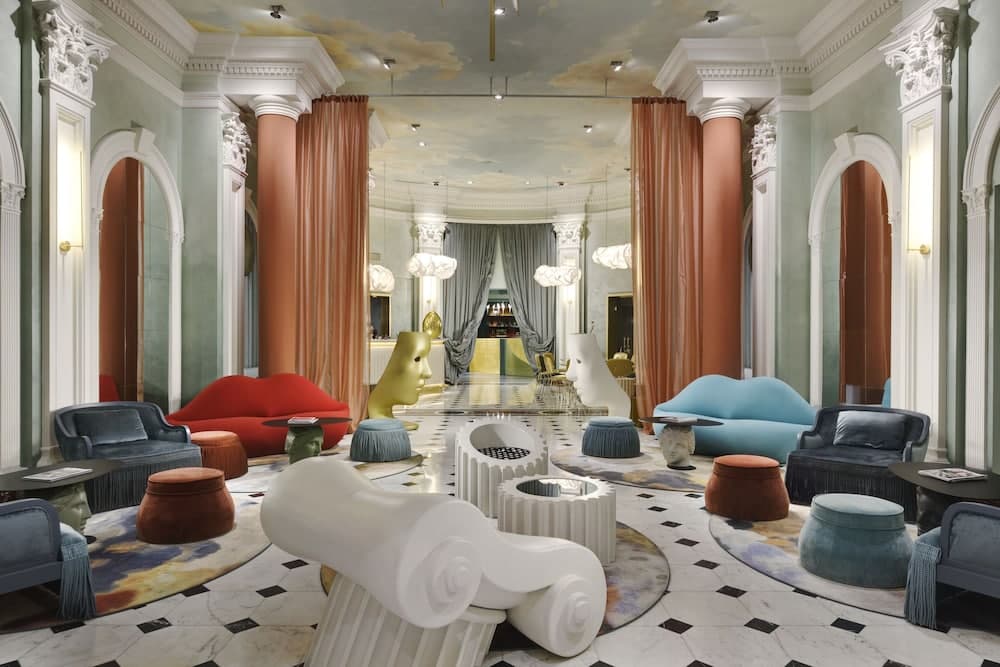 An opulent hotel lobby features a luxurious atmosphere with ornate columns, vibrant furniture, artistic sculptures, and a cloud-painted ceiling, ideal for travelers seeking a blend of elegance and modernity in their accommodations.