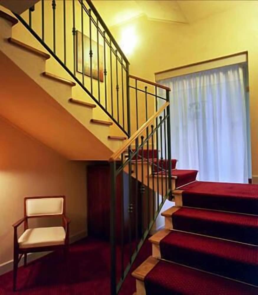 A warmly lit hotel hallway features a staircase with red carpeting and wooden railings, complemented by a cozy chair and soft curtains, ideal for travelers seeking comfort and elegance.