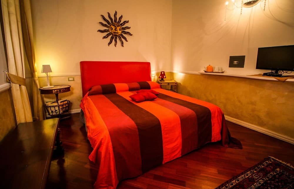 A cozy hotel room features a vibrant red bed adorned with a sunburst wall art, warm lighting, wooden floors, and a flat-screen TV, ideal for travelers seeking comfort and charm.