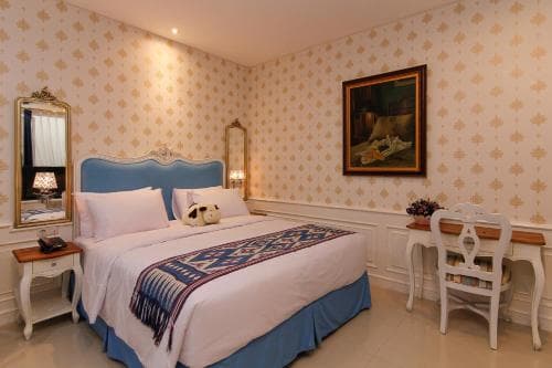 A cozy hotel room features elegant wallpaper, a plush bed adorned with a patterned throw, a classic desk setup, and decorative mirrors, ideal for travelers seeking comfort and charm.