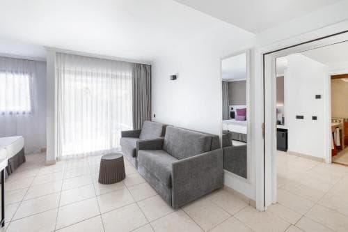 A cozy hotel suite features a modern living area with a gray sofa, tiled floors, sheer curtains, and an open doorway leading to a bedroom, ideal for travelers seeking comfort and style.