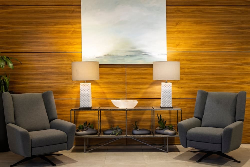 A cozy hotel lobby features modern gray armchairs flanking a sleek console table adorned with white lamps and plants, set against a warm wooden wall with an abstract painting, ideal for travelers seeking a serene and stylish waiting area.