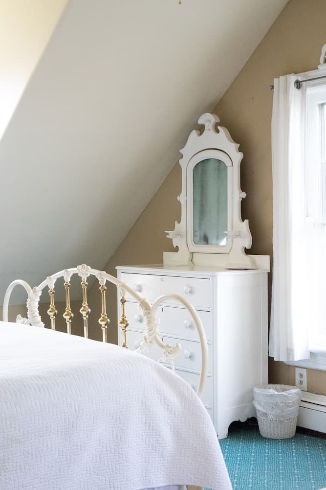 A cozy attic bedroom features a white ornate bed frame, a vintage dresser with a mirror, and soft natural light filtering through a window, creating a serene and charming retreat ideal for a relaxing getaway.