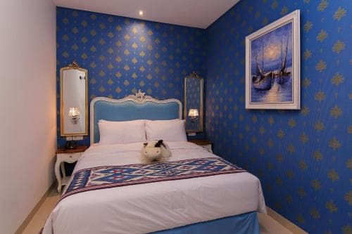 A cozy hotel room features a neatly made bed adorned with a patterned throw blanket, blue walls with gold accents, decorative mirrors, and a serene boat painting, ideal for travelers seeking a tranquil and stylish accommodation.