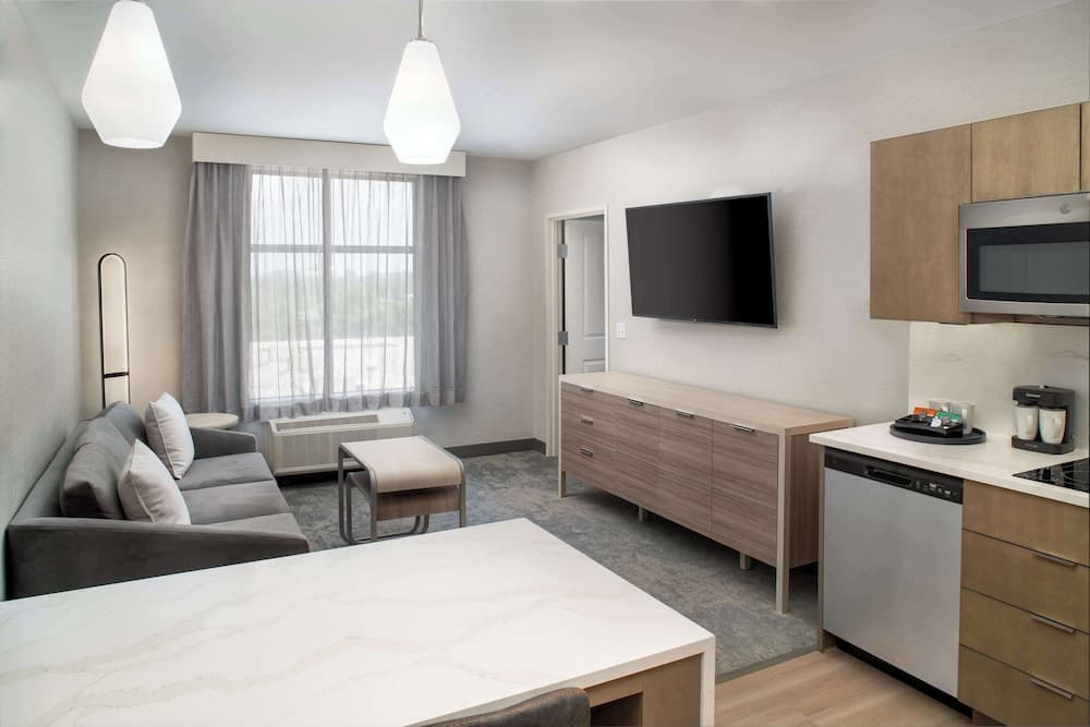 A cozy hotel suite features a modern living area with a plush sofa, sleek kitchenette, flat-screen TV, and large window draped in soft curtains, ideal for travelers seeking comfort and convenience.
