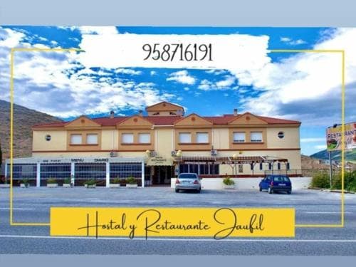 A cozy hostal and restaurant with terracotta roofs and white pillars stands invitingly against a backdrop of clear skies and distant mountains, perfect for travelers seeking a warm meal and comfortable lodging in a serene setting.