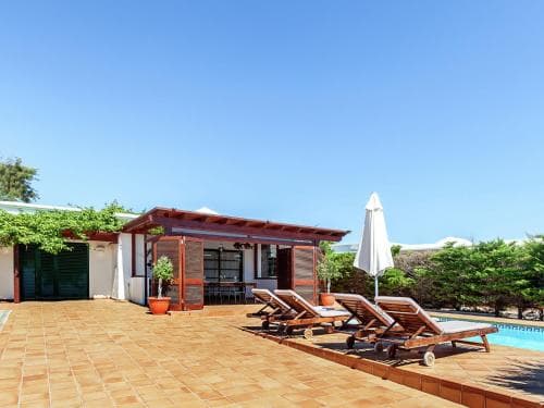 A serene poolside retreat features wooden loungers, a tiled patio, and a cozy cabana under a clear blue sky, ideal for relaxing vacations.