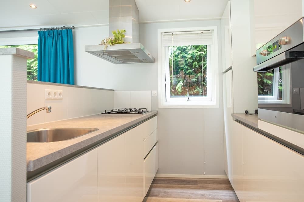 A modern, minimalist kitchen with sleek white cabinets, stainless steel appliances, and a vibrant blue curtain frames a serene view of lush greenery, ideal for travelers seeking a cozy and stylish rental space.