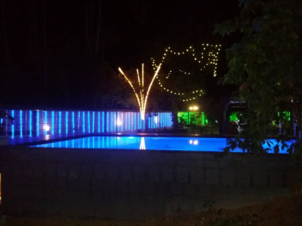 A serene nighttime scene features a glowing blue-lit swimming pool surrounded by festive string lights and trees, ideal for a tranquil evening retreat or romantic getaway.