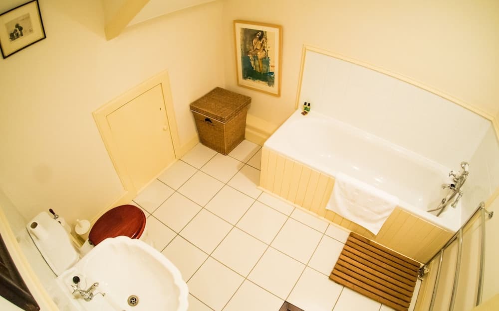 A cozy bathroom with tiled floors, a bathtub adorned with a towel, a wooden toilet seat, framed artwork on the walls, and a wicker basket, ideal for travelers seeking comfort and charm in their accommodations.