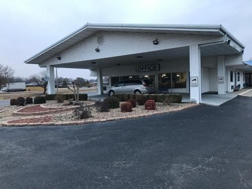 A quiet car dealership office with a covered parking area, landscaped shrubs, and a parked vehicle sets a calm business atmosphere ideal for travelers seeking automotive services or sales.