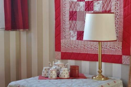 A cozy bedroom corner features a vibrant red quilted wall hanging, a gold lamp on a patterned tablecloth, and charming ceramic teapots, ideal for travelers seeking a warm and inviting accommodation ambiance.