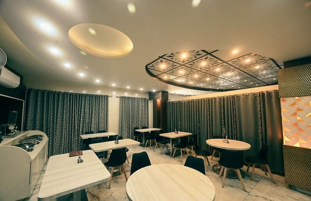 A cozy restaurant interior features modern white tables, black chairs, elegant curtains, and decorative ceiling lights, creating an inviting atmosphere ideal for dining and social gatherings.