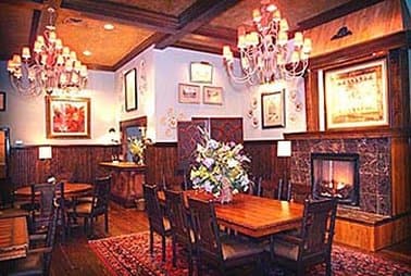 A cozy restaurant interior features warm lighting from elegant chandeliers, wooden tables set for dining, a fireplace adding ambiance, and floral arrangements enhancing the inviting atmosphere, perfect for travelers seeking a charming dining experience.