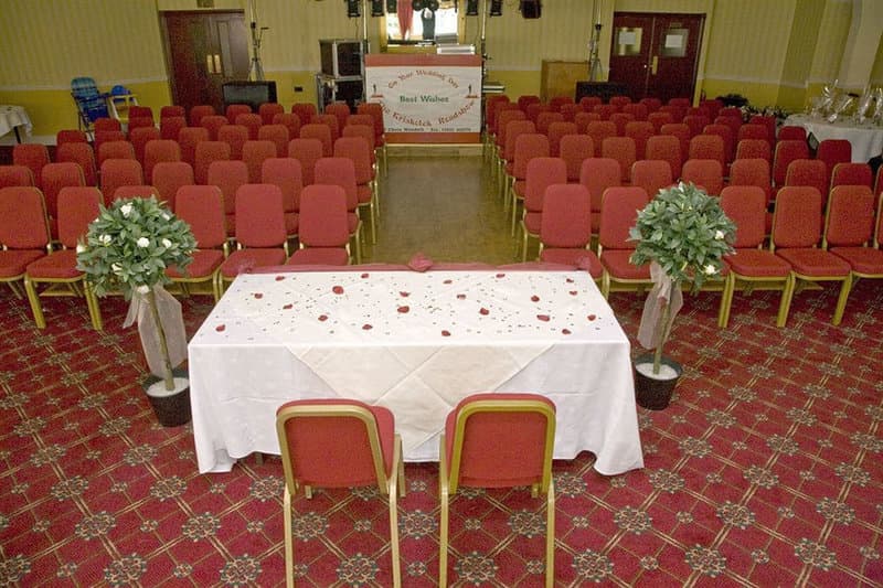 A warmly lit banquet hall set up for a wedding ceremony features rows of red chairs, a central table adorned with white linens and rose petals, potted plants with bows, and a festive atmosphere ideal for intimate celebrations.