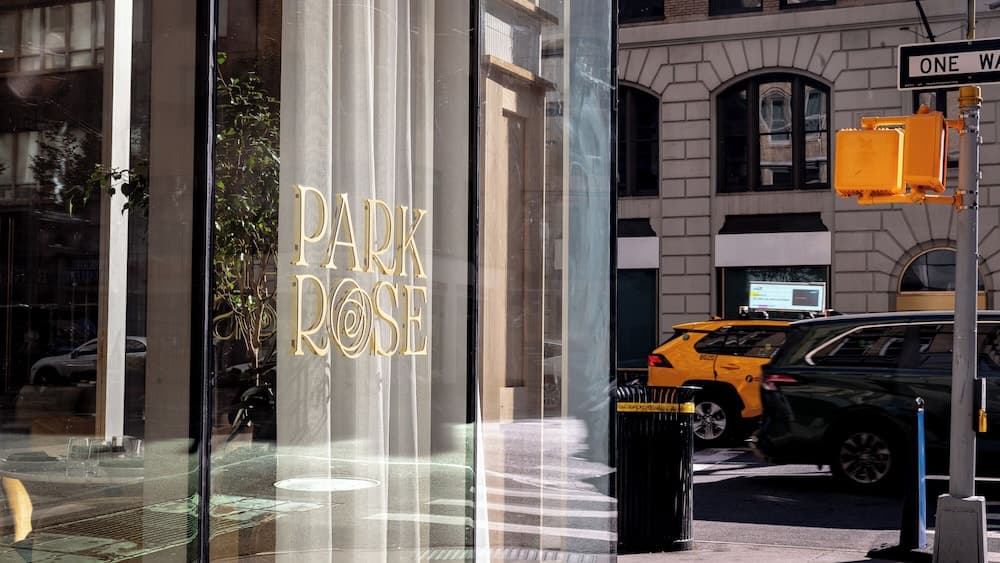 A chic urban street scene features the elegant "Park Rose" signage through a glass storefront, complemented by a bustling sidewalk, classic architecture, and parked cars, ideal for city explorers seeking upscale dining or shopping experiences.