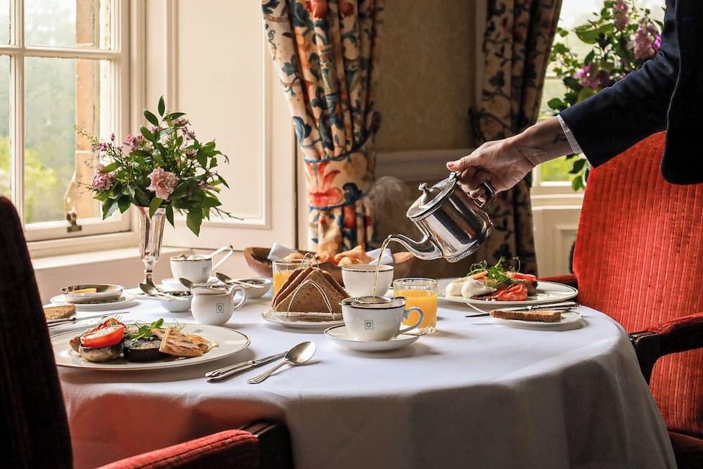 A serene breakfast scene unfolds in an elegant dining room, where a server pours tea onto a table adorned with fresh flowers, colorful curtains, and a variety of appetizing dishes, perfect for travelers seeking a cozy and luxurious morning experience.