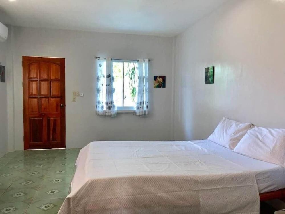 A cozy bedroom with a neatly made bed, patterned green floor tiles, wooden door, and floral curtains framing a window view of greenery, ideal for travelers seeking a tranquil and simple accommodation.