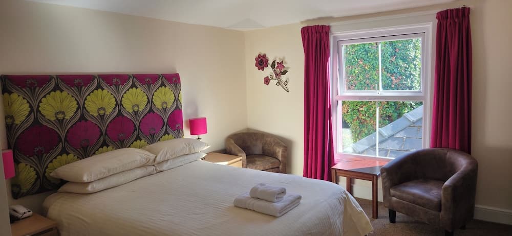 A cozy hotel room features a neatly made bed with a vibrant floral headboard, plush armchairs by a window with lush greenery outside, creating a serene and inviting atmosphere ideal for travelers seeking relaxation.