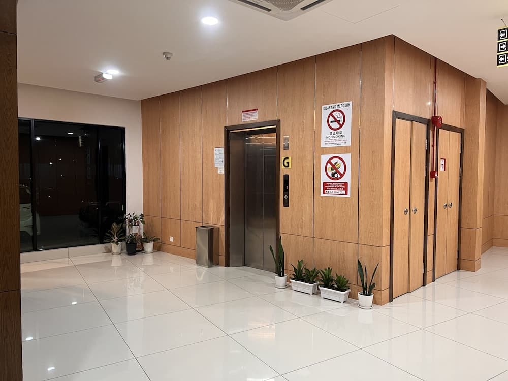 A modern hotel lobby features a sleek elevator, wooden paneling, no-smoking signs, potted plants, and reflective white tiles, ideal for travelers seeking a clean and organized accommodation entrance.
