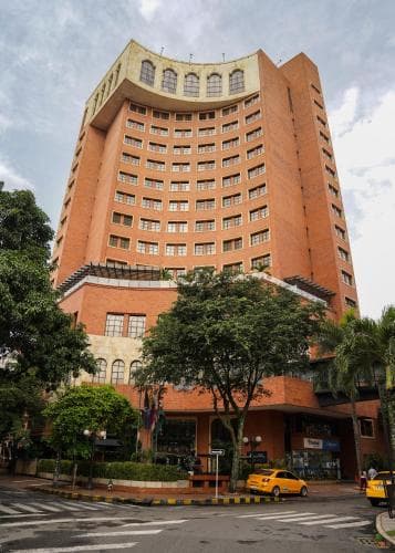 A tall brick hotel stands prominently amidst lush greenery in an urban setting, featuring a yellow taxi parked nearby, ideal for travelers seeking accommodation in a vibrant city atmosphere.