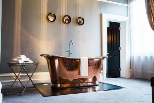 A luxurious bathroom features a gleaming copper bathtub as the centerpiece, complemented by elegant wall mirrors and soft lighting, creating an inviting and serene atmosphere ideal for a high-end hotel stay or spa retreat.