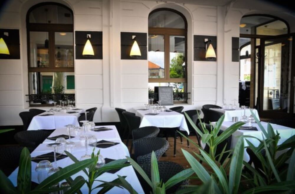 An inviting outdoor dining area with white tablecloths, black chairs, and lush greenery framed by arched windows and warm sconces, perfect for travelers seeking a cozy yet elegant restaurant experience.