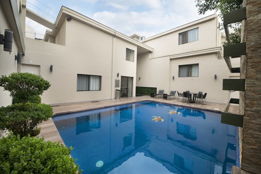 A serene courtyard with a sparkling blue pool surrounded by modern beige walls, lush greenery, and outdoor seating, ideal for a relaxing vacation retreat.