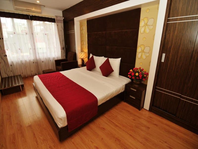 A cozy hotel room features a neatly made bed with red accents, wooden flooring, floral wallpaper, and a bright window, ideal for travelers seeking comfort and style.
