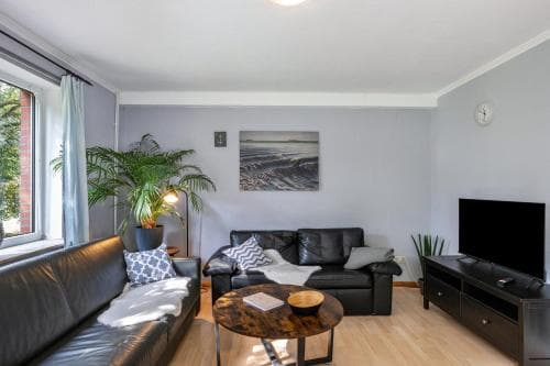 A cozy living room with modern furnishings, including leather sofas, a wooden coffee table, and a large window with greenery outside, creates an inviting atmosphere ideal for travelers seeking comfort in a vacation rental.