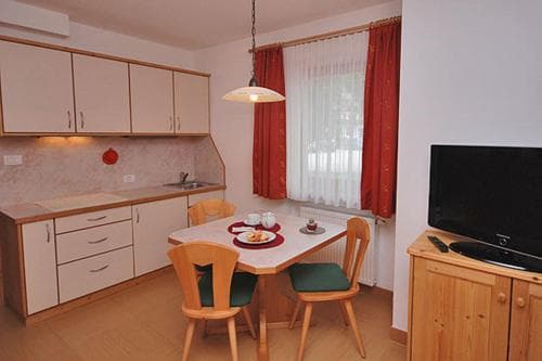A cozy apartment kitchenette features wooden furniture, a dining table set for two by a window with red curtains, and a modern flat-screen TV, ideal for travelers seeking comfort in a compact living space.