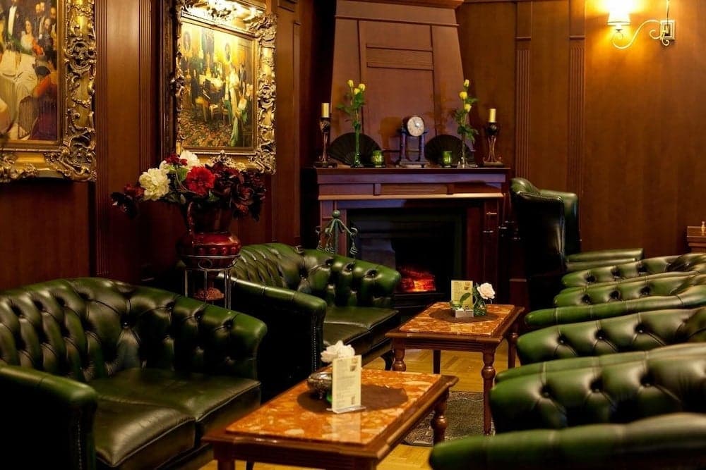 A cozy lounge with plush green leather armchairs, a warm fireplace, ornate paintings, and elegant decor invites travelers to relax in a sophisticated and inviting atmosphere.