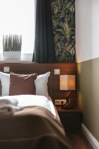 A cozy hotel room features a neatly made bed beside a wooden nightstand with a lamp, set against a backdrop of leaf-patterned wallpaper and a window with greenery outside, ideal for travelers seeking comfort and relaxation.