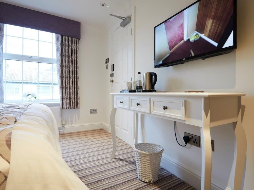 A cozy hotel room features a neatly made bed, a white console table with a kettle and coffee supplies, a wall-mounted TV, and a bright window with patterned curtains, ideal for travelers seeking comfort and convenience.