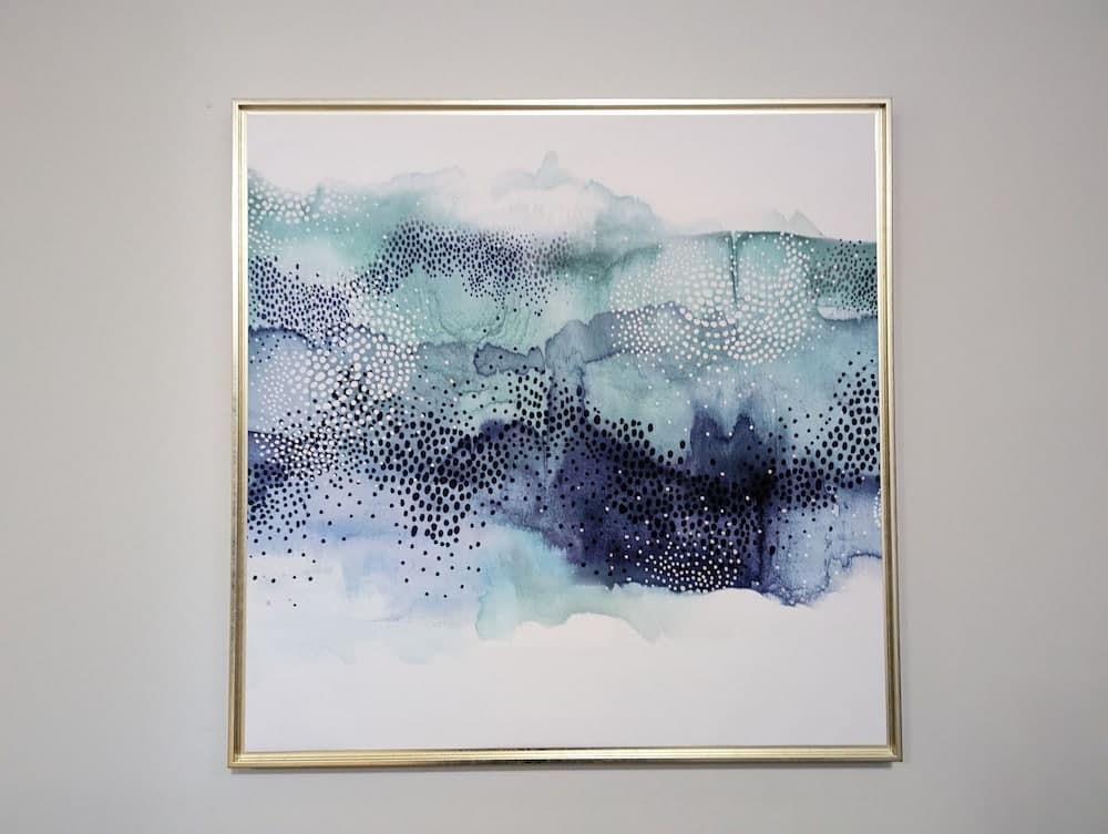 A serene abstract painting featuring blue and white hues with dotted textures hangs in a minimalist indoor setting, ideal for travelers seeking modern art inspiration or home decor ideas.