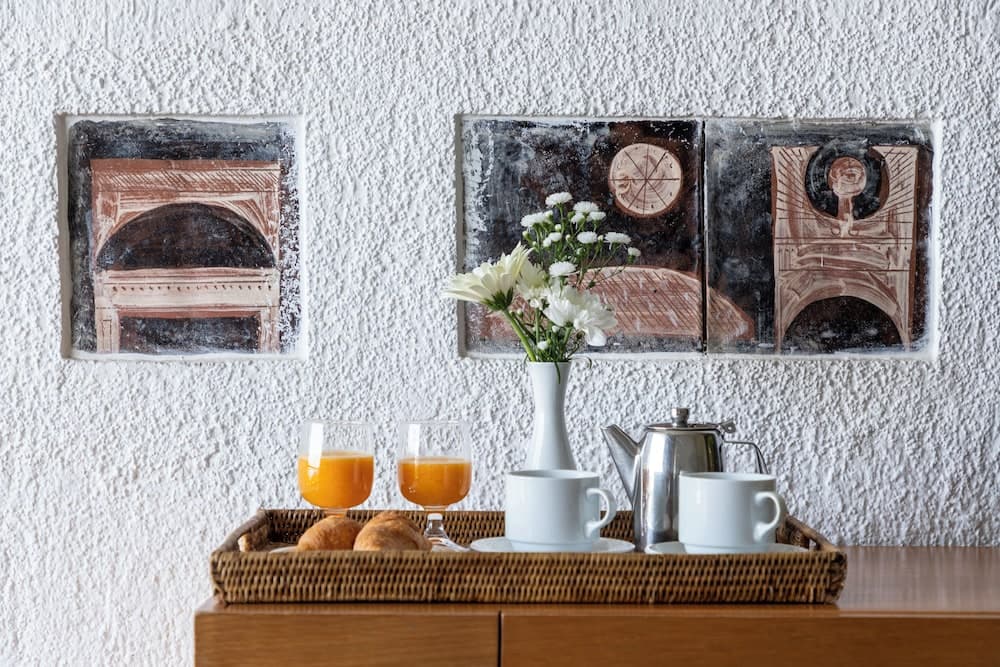 A cozy hotel room breakfast setup features a wooden tray with orange juice, croissants, tea cups, and a vase of white flowers against a textured wall adorned with artistic tiles, ideal for travelers seeking a serene morning experience.