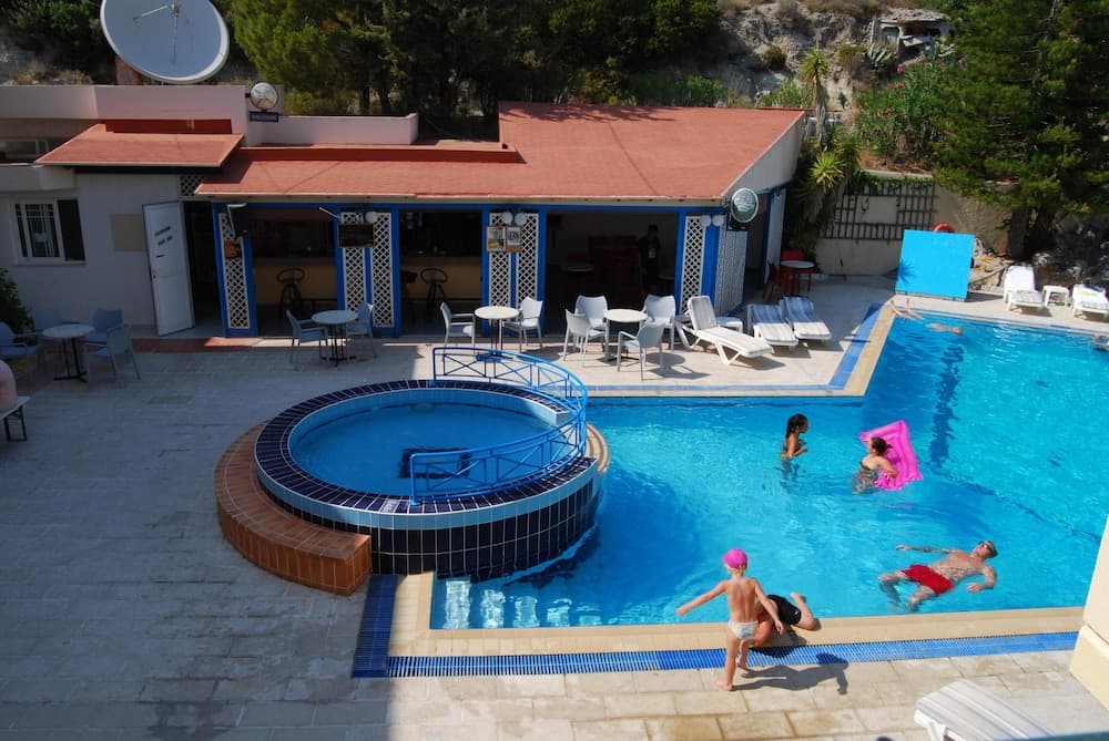 A vibrant resort scene features a large swimming pool with a circular hot tub, surrounded by lounge chairs and a shaded café area, ideal for family vacations in sunny destinations.
