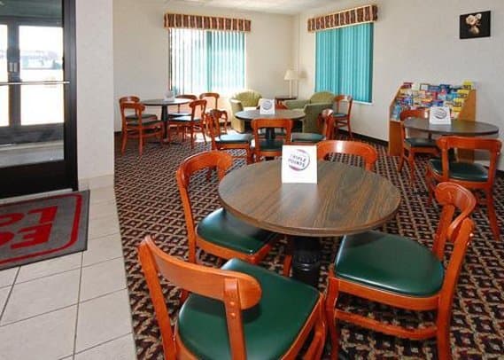A cozy hotel lounge features round tables with green cushioned chairs, patterned carpeting, and bright windows, creating a welcoming atmosphere ideal for travelers seeking a comfortable spot to relax or dine.