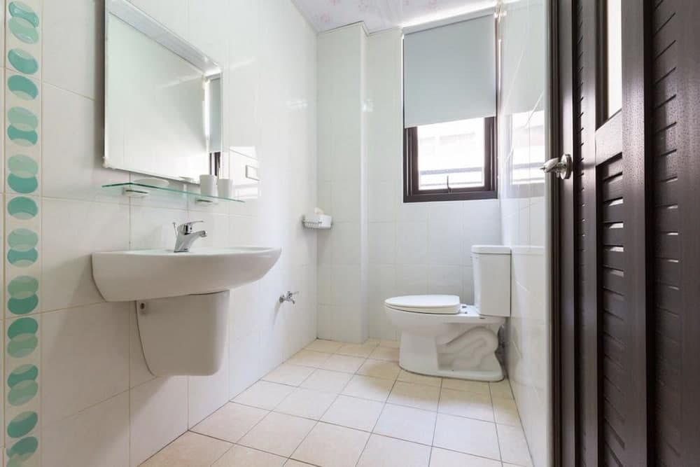 A clean, compact bathroom with white tiles, a sink, toilet, mirror, and window offers a serene space ideal for travelers seeking a simple yet functional accommodation.